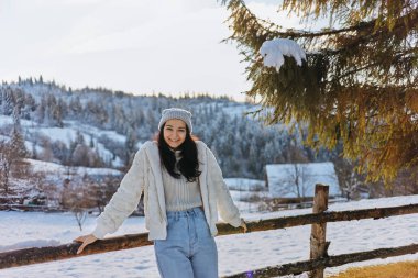 Sıcak kışlık giysiler içinde neşeli bir kadın ahşap bir çite yaslanmış, güneşli kırsalın tadını çıkarıyor. Yaşam tarzı, doğa, tatil veya seyahatle ilgili kavramlar için ideal. 