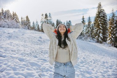 Sıcak kışlık giysiler içinde neşeli genç bir kadın güneşli, karlı bir tarlada duruyor, kolları rahatlamış, güzel bir ormanla çevrili. Yaşam tarzı, doğa ya da mevsimlik temalar için ideal. 