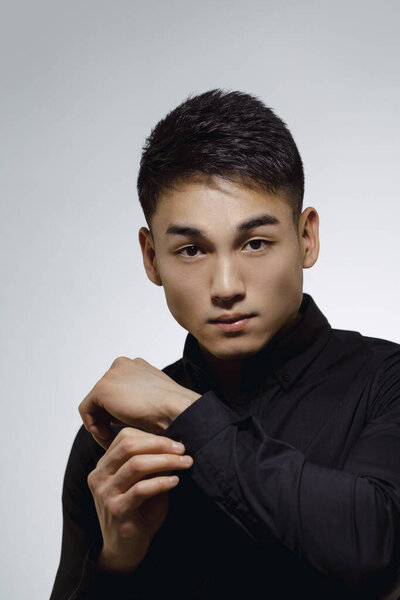 Portrait of a confident young Asian man adjusting his shirt cuff in a studio setting. The serious expression and modern black shirt convey professionalism and style, ideal for business and fashion