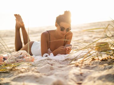  A young woman in a white swimsuit and sunglasses lying on a sandy beach, using her smartphone. Captures summer relaxation, trendy lifestyle, and serene moments by the seaside. clipart