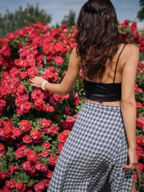 Siyah ekin bluzlu ve uzun damalı etekli şık bir kadın çiçek açan kırmızı güllerle dolu bir bahçenin tadını çıkarıyor. Doğa, moda ve yaşam tarzı temalı içerik için ideal olan bu yüksek kaliteli fotoğraf,