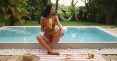 Bikinili neşeli bir kadın, elinde bir şişe taze meyve suyu olan lüks bir havuzda dinleniyor. Tropikal yeşillik ve tatil atmosferiyle çevrili bu sahne rahatlama ve mutluluk yayar.. 