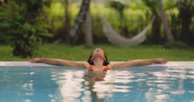 Kırmızı bikinili sakin bir kadın kollarını açmış tropikal bir havuza uzanıyor ve bir an rahatlıyor. Bereketli yeşil zemin ve hamak huzur ve tatil atmosferine ekleniyor..