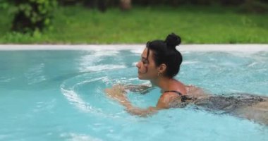 Açık havuzda zarif bir şekilde yüzen siyah mayo giymiş bir kadının sakin bir sahnesi. Ağır çekimde yakalanan bu görüntüler gevşemeyi, zarafeti ve tropikal güzelliği vurguluyor..