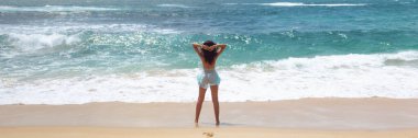 Arkadaki görüntüde kumlu bir sahilde çıplak ayakla duran bir kadın turkuaz okyanus dalgalarına bakıyor. Özgürlük, rahatlama ve doğa ile bağlantı anı yakalar, seyahat ve yaşam tarzı teması için idealdir..
