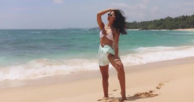 Güneşli bir tropikal plajda bikini ve güneş gözlüğü takan kendine güvenen genç bir kadın. Yaz, seyahat, moda ve plaj yaşam tarzı temaları reklamcılık ya da promosyonlar için mükemmel. 