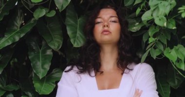 Beyaz elbiseli çarpıcı esmer bir kadın kolları çapraz duran, özgüven ve özgüven saçan gür tropikal yaprakların arasında duruyor. Cesur moda, güçlendirme ve yaşam tarzı projeleri için ideal.. 