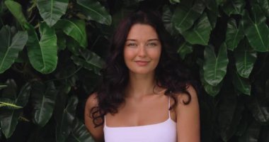 Koyu kıvırcık saçlı, beyaz bluzlu ve canlı tropikal yapraklara karşı duran çarpıcı bir genç kadın. Güzellik, kaplıca, cilt bakımı ya da eko-yaşam tarzı kavramları için mükemmel.. 