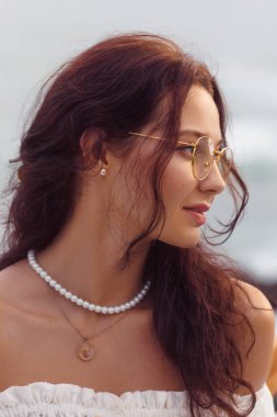 Açık hava ortamında yuvarlak gözlüklü ve inci kolyeli zarif bir esmer kadın profili. Görüntü onun doğal güzelliğini vurguluyor, onu moda, yaşam tarzı ve...