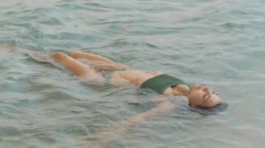 Yeşil bir bikini giyerken sakin okyanus dalgalarında süzülen bir kadının sakin bir sahnesi. Rahatlamanın, yaz atmosferinin ve sükunetin mükemmel tasviri.. 
