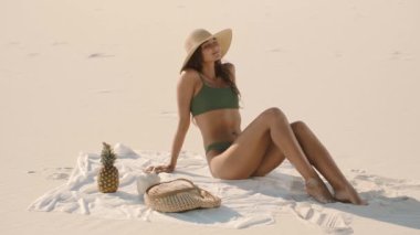Yeşil bikinili ve güneş şapkalı genç bir kadın kumsalda kumsalda bir havluya yaslanıyor. Etrafı ananas ve yaz esaslarıyla çevrili. Tatil, seyahat ve tropikal yaşam tarzı temaları için ideal.