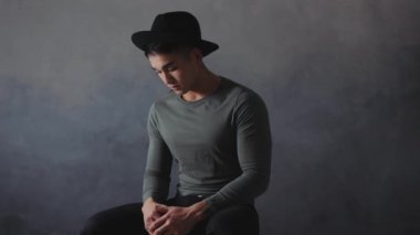 Siyah şapkalı ve gri tişörtlü dalgın bir Asyalı, koyu arkaplanlı, minimalist bir stüdyoda tek başına oturuyor. Ruh hali kasvetli, hüzün, yalnızlık temalarını yansıtıyor.
