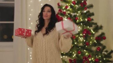 Noel Baba şapkalı ve rahat süveterli mutlu bir kadın Noel hediyeleriyle parıldayan ışıklarla süslenmiş güzel bir ağacın önünde dans ediyor. Tatil promosyonları ve şenlikli pazarlama için mükemmel..