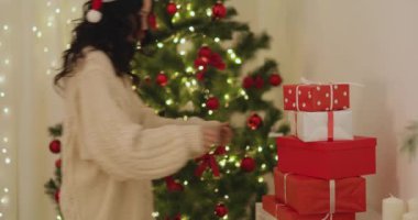 Noel Baba şapkası takan ve rahat süveter giyen bir kadın, parlak ışıkları olan güzel dekore edilmiş bir ağacın yanındaki kırmızı ve beyaz Noel hediyelerini kaldırıyor. Tatil promosyonları ve şenlik için ideal
