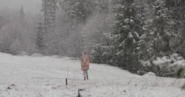 Sıcak palto ve kış kıyafetleri giymiş bir kadın karla kaplı sakin bir ormanda yürüyor. Huzurlu kış sahneleri yağan kar, yemyeşil ağaçlar ve doğal güzellikleri gözler önüne seriyor..