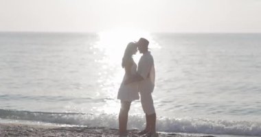 Sevgi dolu bir çift gün batımında sakin bir plajda romantik bir öpücük paylaşır. Altın güneş ışığı, sakin okyanus dalgalarını yansıtır. Aşkı simgeleyen barışçıl ve samimi bir atmosfer yaratır.