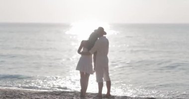 Beyaz elbiseli romantik bir çift gün batımında parıldayan okyanus dalgalarını izlerken kucaklaşır. Huzurlu sahne aşkı, samimiyeti ve kıyı manzarasının güzelliğini temsil eder..