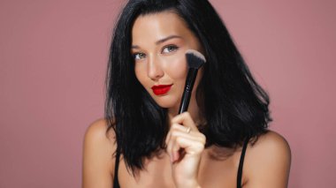 Çarpıcı kırmızı dudakları ve siyah saçları olan zarif bir kadın pembe arka planda yanağına yakın bir makyaj fırçası tutuyor. Güzellik dersleri, kozmetik aletleri reklamları ve makyaj sanatçıları için mükemmel.