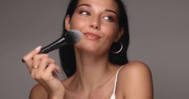 Kendine güvenen bir kadının makyaj fırçasıyla doğal güzelliğini arttırdığı yakın plan. Zarafet ve stile odaklı kozmetik, güzellik ürünleri veya makyaj derslerini tanıtmak için ideal. 