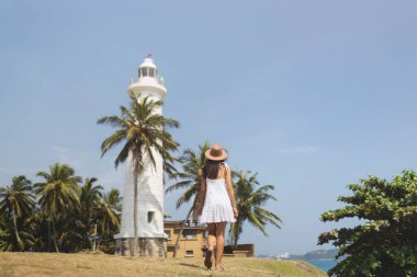 Beyaz elbiseli ve şapkalı bir kadın Galle Fort, Sri Lanka 'daki bir deniz fenerine doğru yürüyor. Tropikal palmiye ağaçları ve berrak mavi gökyüzü turizm için mükemmel ve huzurlu bir seyahat konsepti oluşturur.