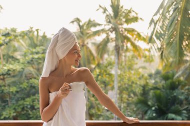 Kafasında havluyla gülümseyen bir kadın, elinde bir kahve fincanı, bir balkonda dikiliyor, yemyeşil bir yeşil ile çevrili. Rahatlama, kişisel bakım ve sabah sağlığı kavramları için idealdir.. 