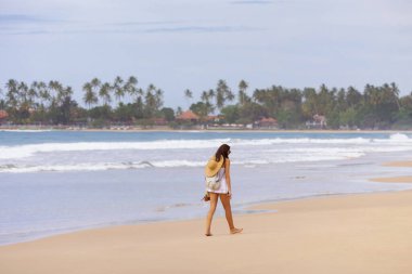 Dickwella, Sri Lanka 'daki sakin plajda palmiye ağaçları ve arka planda deniz kıyısında bir tatil köyü olan günlük kıyafetleriyle gezinen bir kadın. Seyahat, rahatlama ve tropikal konular için mükemmel.