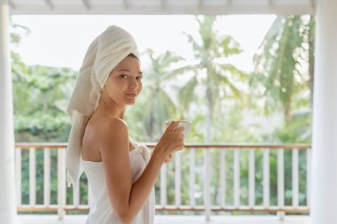 Kafasına ve vücuduna havlu sarılmış bir kadın, tropik yeşilliğe tepeden bakan bir balkonda sabah kahvesinin tadını çıkarıyor. Rahatlama ve sabah rutini konsepti. Açık hava fotoğrafçılığı
