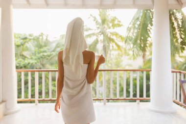 Havluya sarılmış bir kadın, tropik bir manzaraya tepeden bakan bir balkonda duruyor. Huzurlu sabah atmosferinin tadını çıkarıyor. Huzur ortamı gevşemeyi, kendine bakmayı ve tropikal yaşam lüksünü teşvik eder.