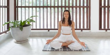 Huzurlu bir kadın nilüfer çiçeğinin içinde yoga minderinin üzerinde oturur, parlak ve huzurlu bir iç mekanda meditasyon yapar. Beyazlar içinde, farkındalığı ve sükuneti kucaklıyor, sakin bir sağlık ortamı yaratıyor..