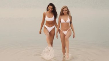 Eşleşen beyaz bikinili iki kadın, sakin ve sığ suda yürürken su sıçratıp el ele tutuşuyorlar. Sahne neşe, dostluk ve yaz rahatlığı yayar. Yaşam tarzı ve plaj için mükemmeldir.