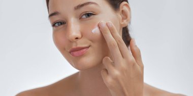 Yüzüne nazikçe cilt kremi süren parlak ciltli bir kadın. Yumuşak ışıklandırma ve kusursuz ten bu görüntüyü güzellik, cilt bakımı ve sağlık terfileri için mükemmel kılıyor.