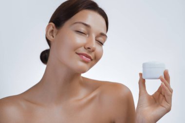 Gözleri kapalı, elinde bir kavanoz cilt kremi tutan ve nazikçe gülümseyen, ışıl ışıl cildini gösteren dingin genç bir kadın. Güzellik ürünleri reklamları, cilt bakımları, promosyonlar, sağlık ve kişisel bakım için harika.