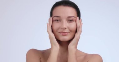 Yüzüne hafifçe dokunurken gülümseyen pürüzsüz, parlak bir teni olan genç bir kadın. Yüz gerdirme, estetik ameliyat ve cilt bakımı için mükemmel. Yüksek kaliteli stüdyo videosu.