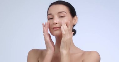 Yüzünde ellerle gülümseyen, kusursuz cildini gösteren güzel bir genç kadın. Cilt bakımı reklamları, doğal güzellik promosyonları ya da markalaşma için mükemmel. Profesyonel stüdyo videosu.