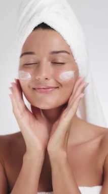 Minimalist bir stüdyoda yüzüne nazikçe cilt kremi sürerek başını havluyla saran gülümseyen bir kadın. Güzellik, sağIık ve cilt bakımı için mükemmel.. 