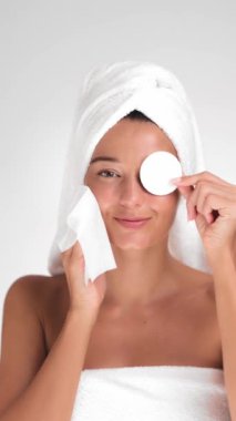 Kafasına havlu sarılmış genç bir kadın yüzünü pamuk pedlerle nazikçe temizliyor, parlak minimalist bir ortamda gülümsüyor. Cilt bakımı, güzellik ve sağlık kavramları için idealdir.. 