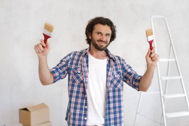 İki boya fırçası tutan, günlük kıyafetli neşeli bir adam, DIY ev yenileme projesine hazır. Ev yenileme, iç tasarım ve yaratıcı projeler için mükemmel bir kavram.. 
