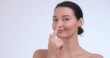 Neşeli bir kadının portresi. Parlak bir teni ve neşeli bir duruşu var. Burnuna bir gülümsemeyle işaret ediyor. Cilt bakımı reklamları, güzellik kampanyaları ve kişisel bakım ürünleri promosyonları için mükemmel.. 