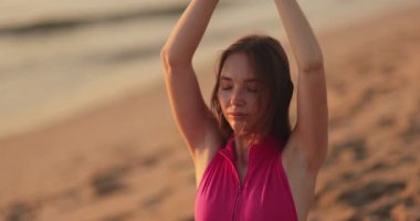 Huzurlu bir kadın gün doğumunda kumsalda meditasyon yapıyor, kolları havada ve gözleri kapalı, farkındalık ve sükuneti kucaklıyor. Açık hava sağlığı, rahatlama ve sağlıklı yaşam için mükemmel.