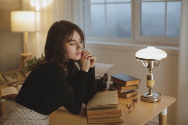 Siyah bluzlu ve ekose etekli şık genç bir kadın ahşap bir masada oturuyor, etrafı kitaplarla çevrili ve parlak bir lambayla, düşünceli bir ifadeyle başını ellerinin üzerinde dinlendiriyor. Yaşam tarzı için ideal