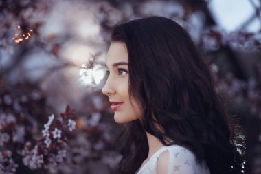 Dalgalı siyah saçlı zarif genç kadın alacakaranlıkta açan kiraz çiçeklerine hafifçe bakıyor. Romantik, çiçekli ve sanatsal yaşam tarzı fotoğrafçılığı için mükemmel.. 
