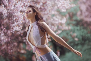 Kolsuz, beyaz bir bluz ve dalgalı bir duruş içinde, çiçek açan kiraz çiçekleriyle çevrili zarif genç bir kadın. Bahar temaları, romantik moda ve sanatsal yaşam tarzı fotoğrafçılığı için mükemmel.. 