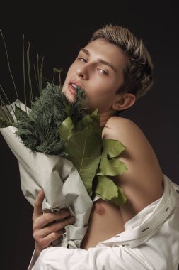 Elinde yemyeşil bir buket tutan, çıplak bir omzu olan, koyu bir arka planda duran genç bir adamın stilize edilmiş portresi. Moda yazıları ve yaratıcı, doğadan esinlenilmiş kampanyalar için ideal..