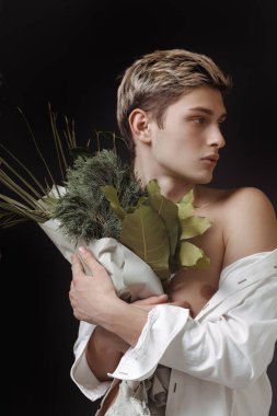 Koyu bir arka planda çıplak omuz ve dramatik ışıklandırma ile bir buket yeşil yaprak tutan genç bir adamın cesur portresi. Moda, sanat ve editör projeleri için mükemmel..