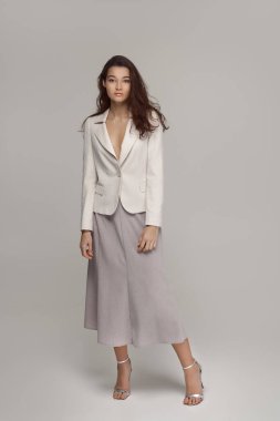 Beyaz ceket giyen, tarafsız etek ve topuklu ayakkabı giyen şık bir kadının tam vücut portresi, minimal bir stüdyo arka planında güvenle poz veriyor. Moda ve yaşam tarzı projeleri için mükemmel..