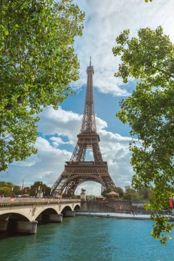 Eiffel Kulesi 'nin göz kamaştırıcı manzarası yemyeşil yeşilliklerle çevrili, sakin bir nehri geçen manzaralı bir köprü. Seyahat, kültür ve doğadan esinlenilmiş Paris görselleri için ideal..