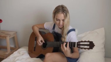 Genç bir kız evde yatağında oturuyor, doğal ışıkta akustik gitar çalıyor. Sahne yaratıcılığı, rahatlığı ve minimalist bir ortamda rahat bir yaşam tarzını ön plana çıkarıyor..