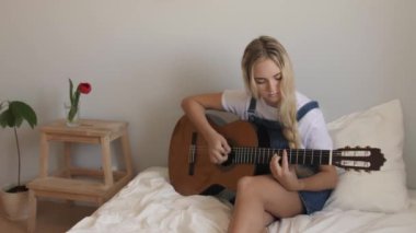 Genç bir kız sıcak bir ev ortamında yatakta otururken akustik gitar çalar. Gençlik, müzik, yaratıcılık ve rahat yaşam tarzı kavramları için mükemmel..