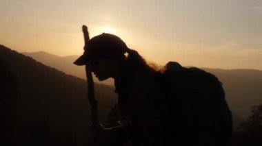 Sırt çantalı ve bastonlu bir kadın yürüyüşçünün el kameralı silueti dağlarda gün batımına karşı hareket ediyor. Macera, doğa ve huzurlu seyahat temaları için mükemmel.