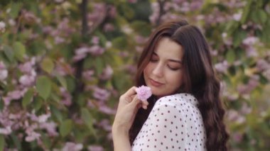 Uzun siyah saçlı, gülümseyen genç bir kadın canlı bir çiçek bahçesinde dururken pembe bir çiçek tutuyor. Sahne zarafeti, doğayı ve ilkbaharın güzelliğini yakalar..